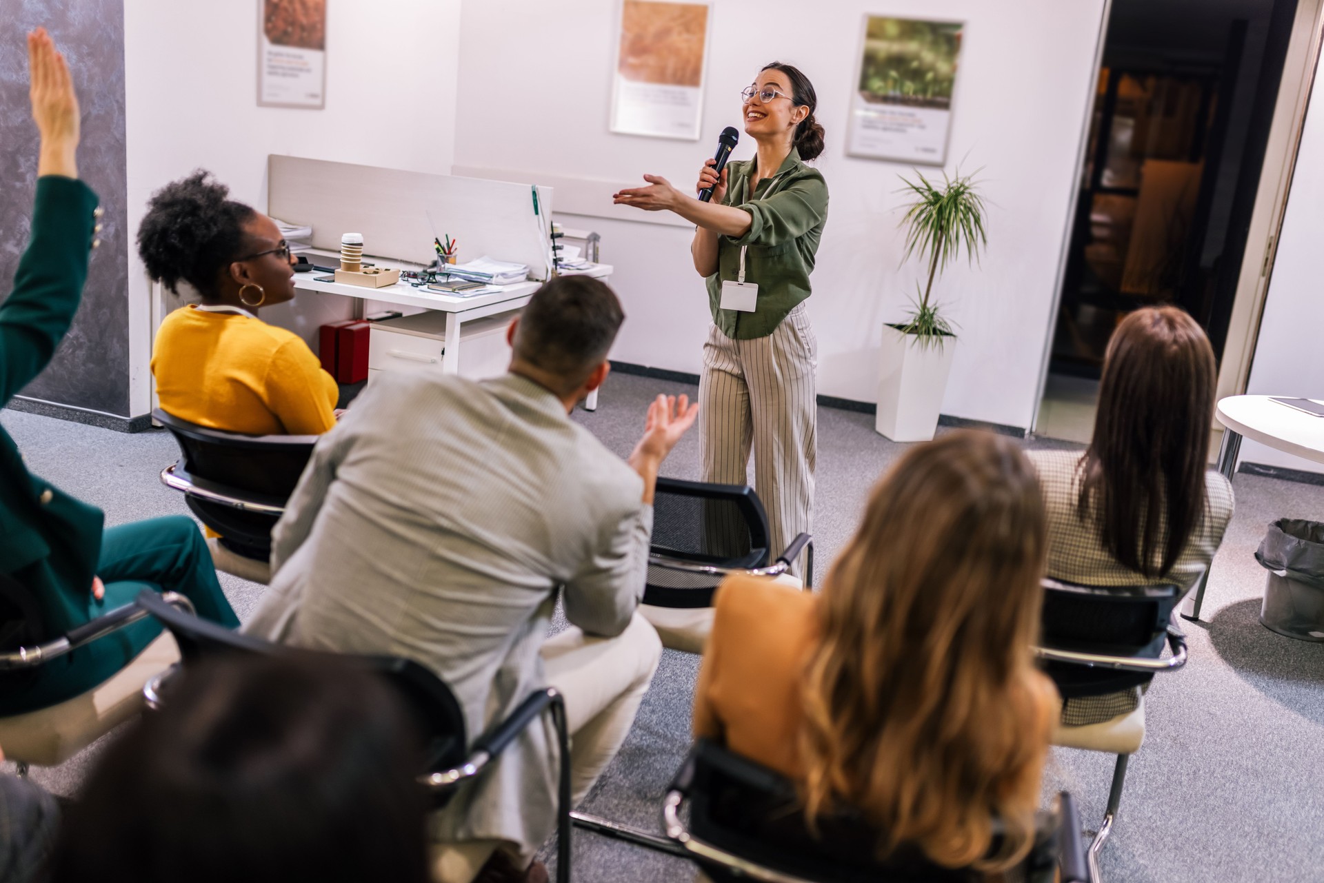 Young bussines woman lecturing her multiethnic colleagues. Having a speach while holding a microphone. Everyone is listening and some are raising their hand.